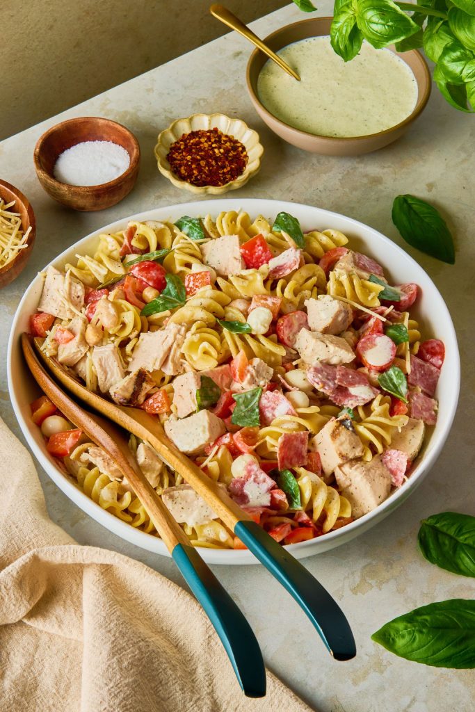 high protein pasta Salad in a bowl with 2 wooden serving spoons on a counter with more ingredients surrounding the bowl