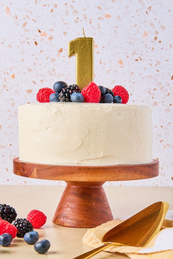 cake frosted on a cake stand topped with fresh berries and a number 1 candle