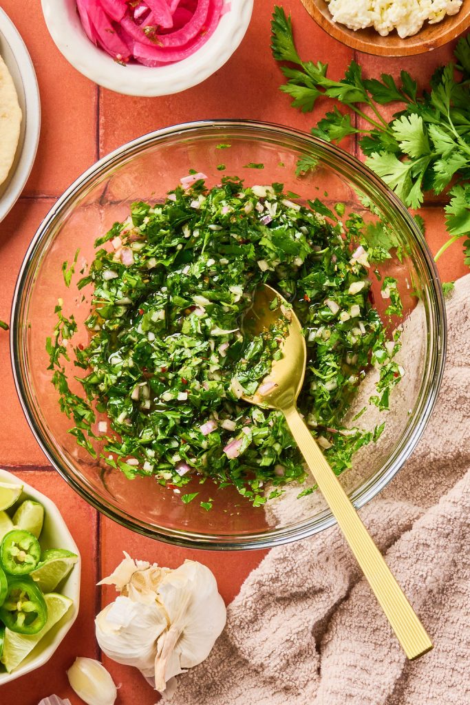 all the chimichurri ingredients mixed together with a gold spoon in a glass bowl