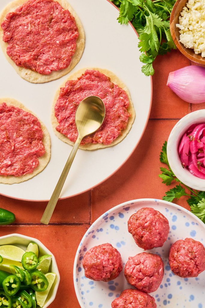 ground beef being smashed into tortillas with a gold spoon
