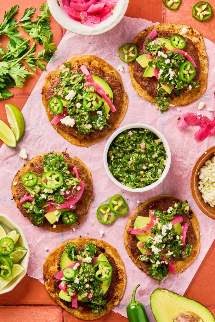 chimichurri smash tacos topped with avocado, jalapeño, and pickled red onions laid out on display with lime wedges