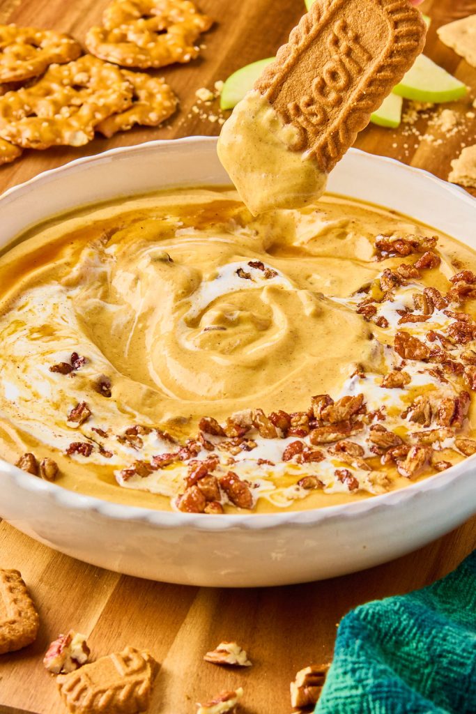 pumpkin dip in a bowl topped with maple pecans and a biscoff cookie being dipped into the dip