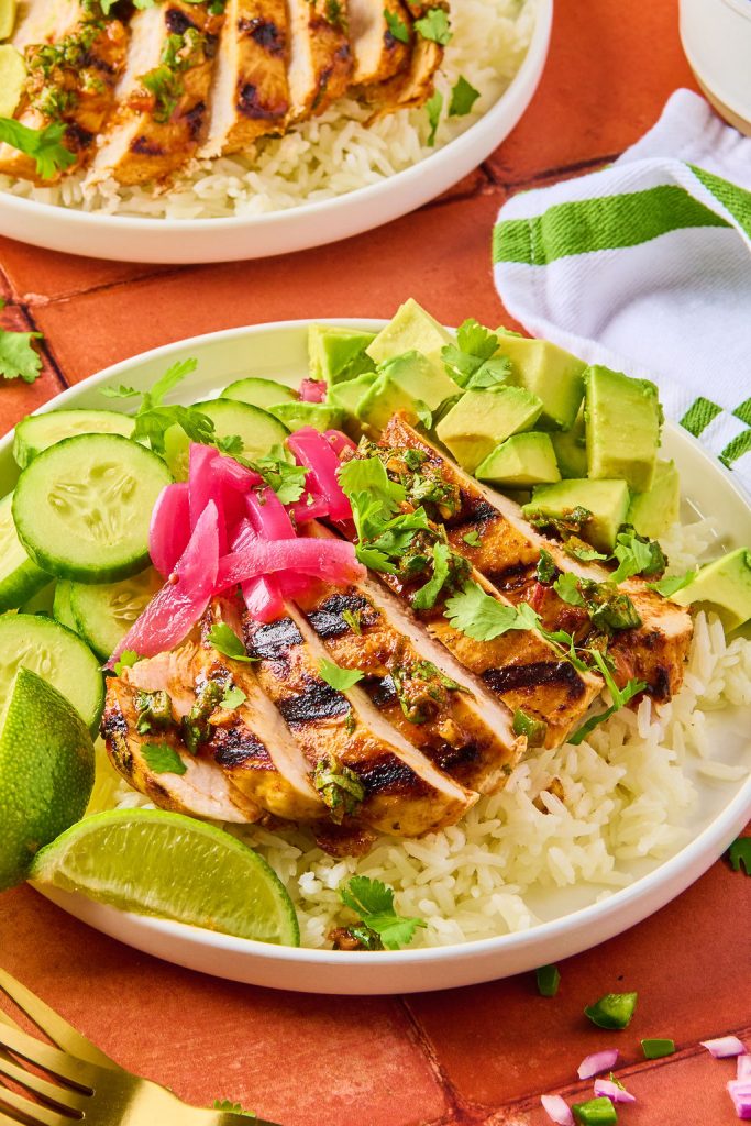 sliced cilantro lime chicken over rice in a bowl, topped with lime wedges, cilantro, red onions, avocado and cucumbers