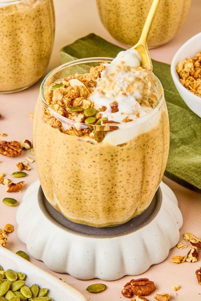 pumpkin pie chia pudding in a glass cup topped with greek yogurt, granola, pumpkin seed and nuts being scooped with a gold spoon