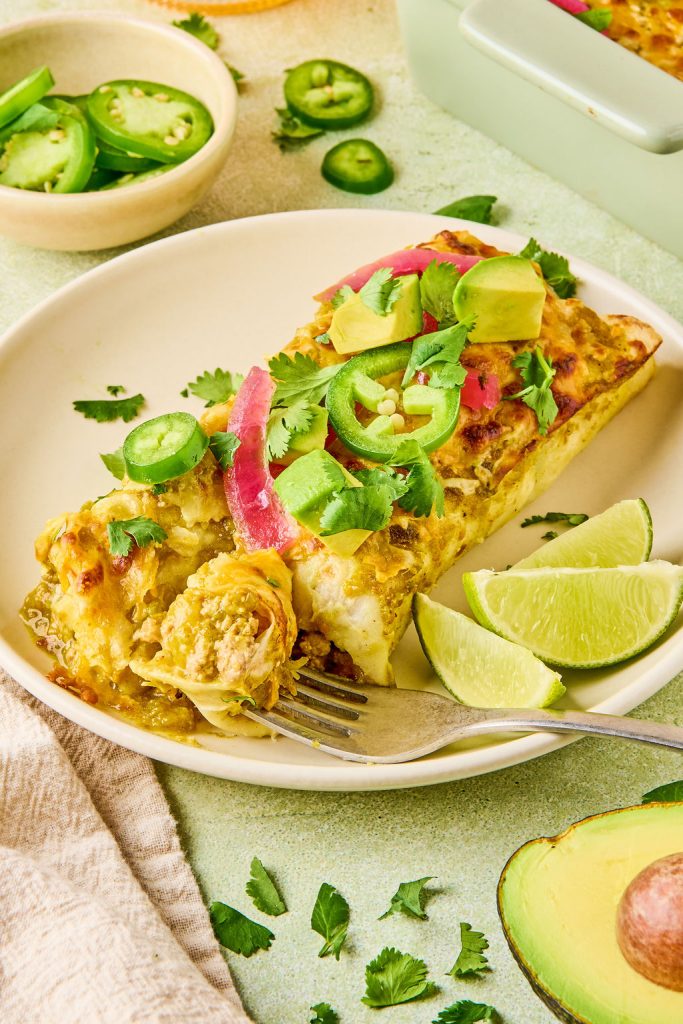 Turkey enchiladas on a plate topped with cilantro, jalapeños, red onion, avocado and lime wedges